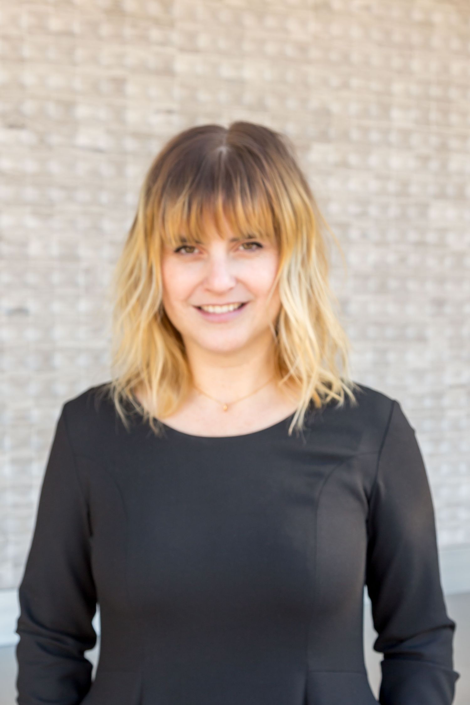 A smiling woman with blonde-tipped shoulder-length hair and bangs, wearing a black dress, poses in front of a light brick wall.