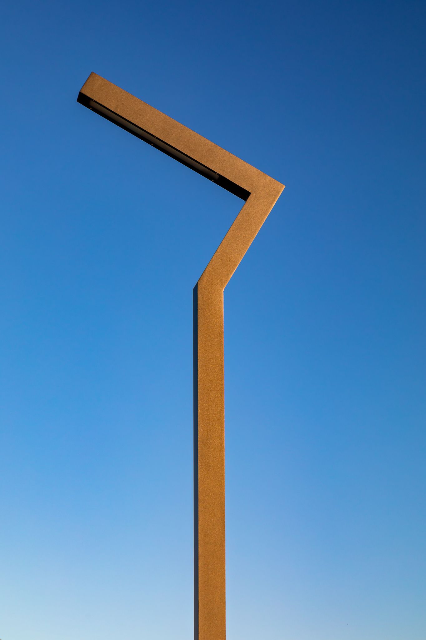 Angular rust-colored steel pole with a bent top arm standing tall against a clear blue sky.
