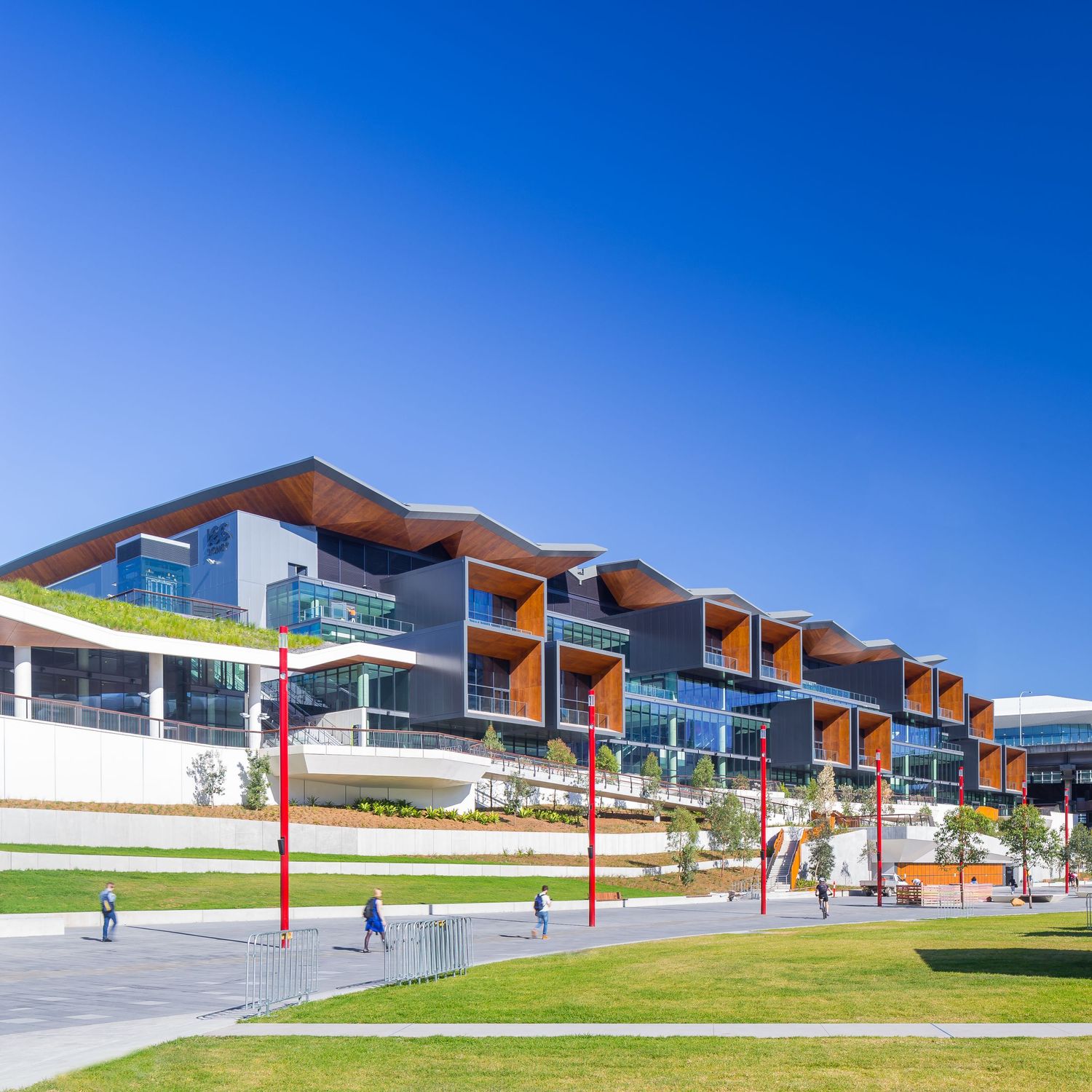 Modern multi-level waterfront building with timber-clad rooflines, glass facades, red poles and landscaped lawns under a clear blue sky.
