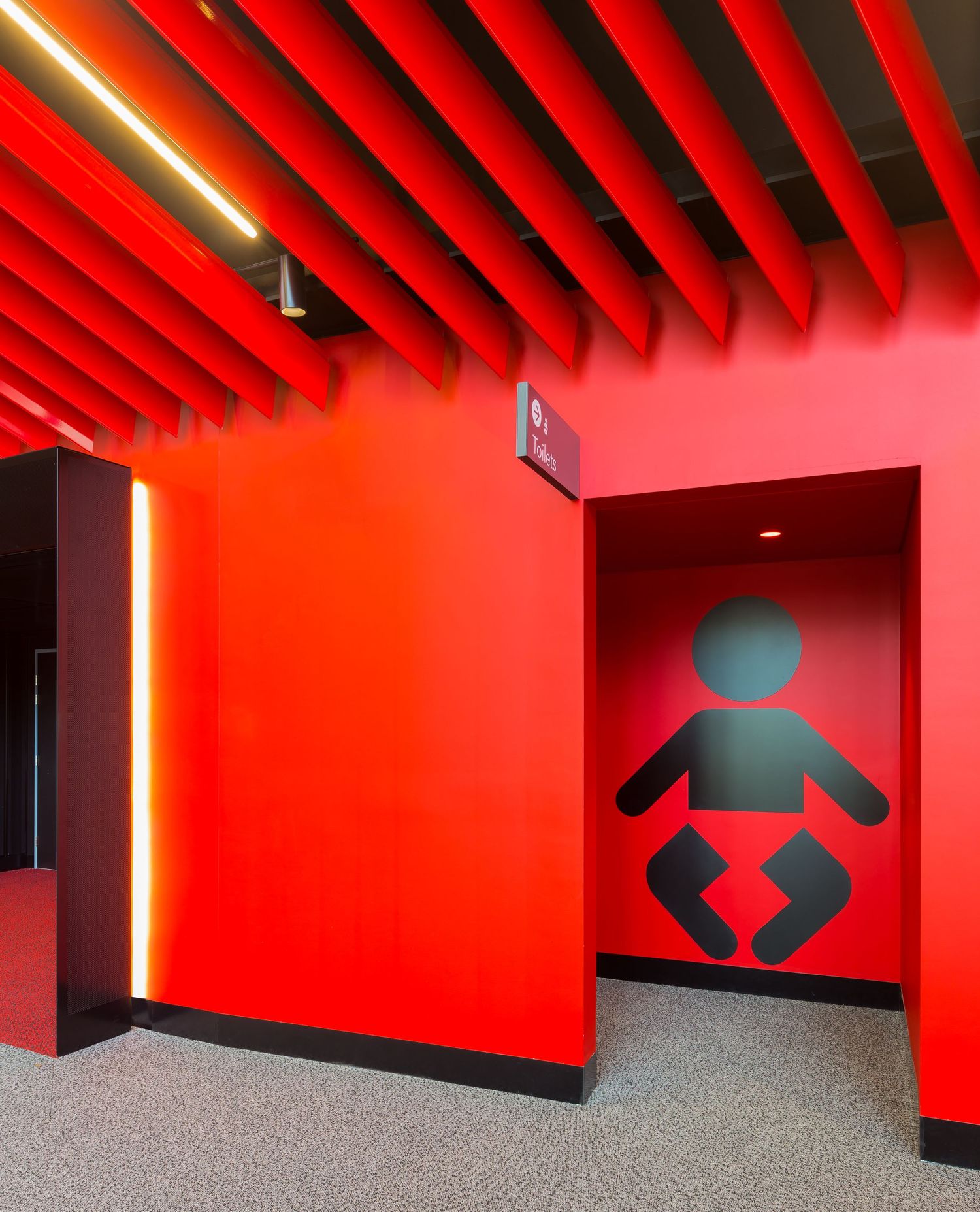 Bright red wall with a black baby-changing symbol at a family room entrance, beneath a red slatted ceiling and warm vertical light strip.