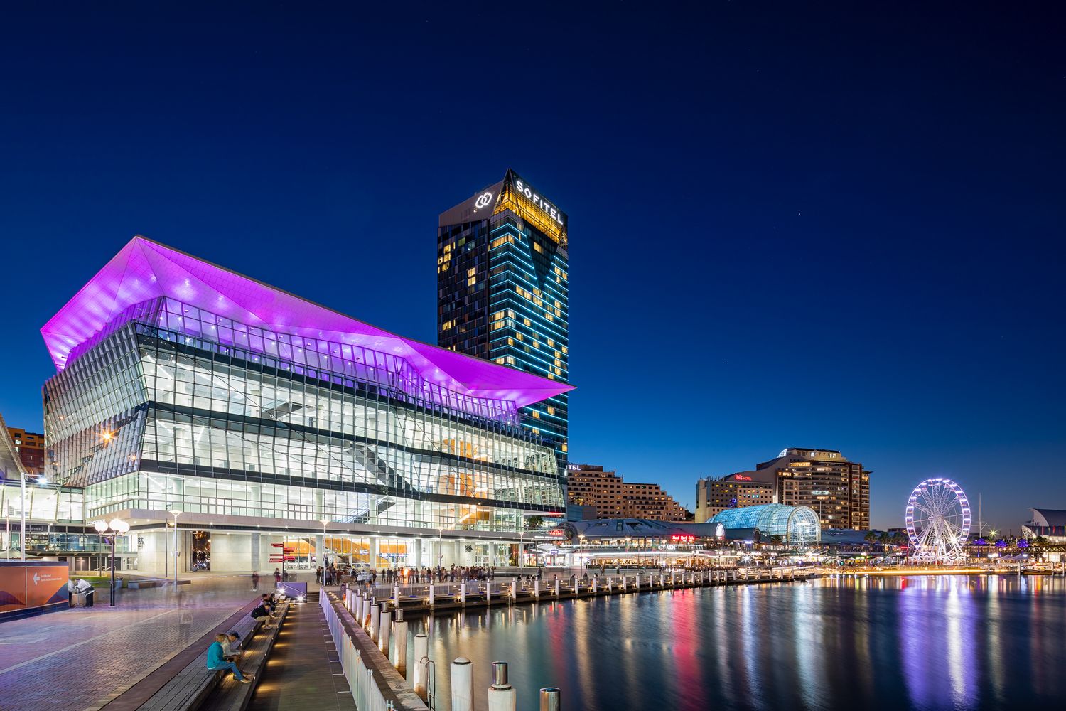 Convention centre glowing purple at night beside Darling Harbour with a hotel tower and ferris wheel.