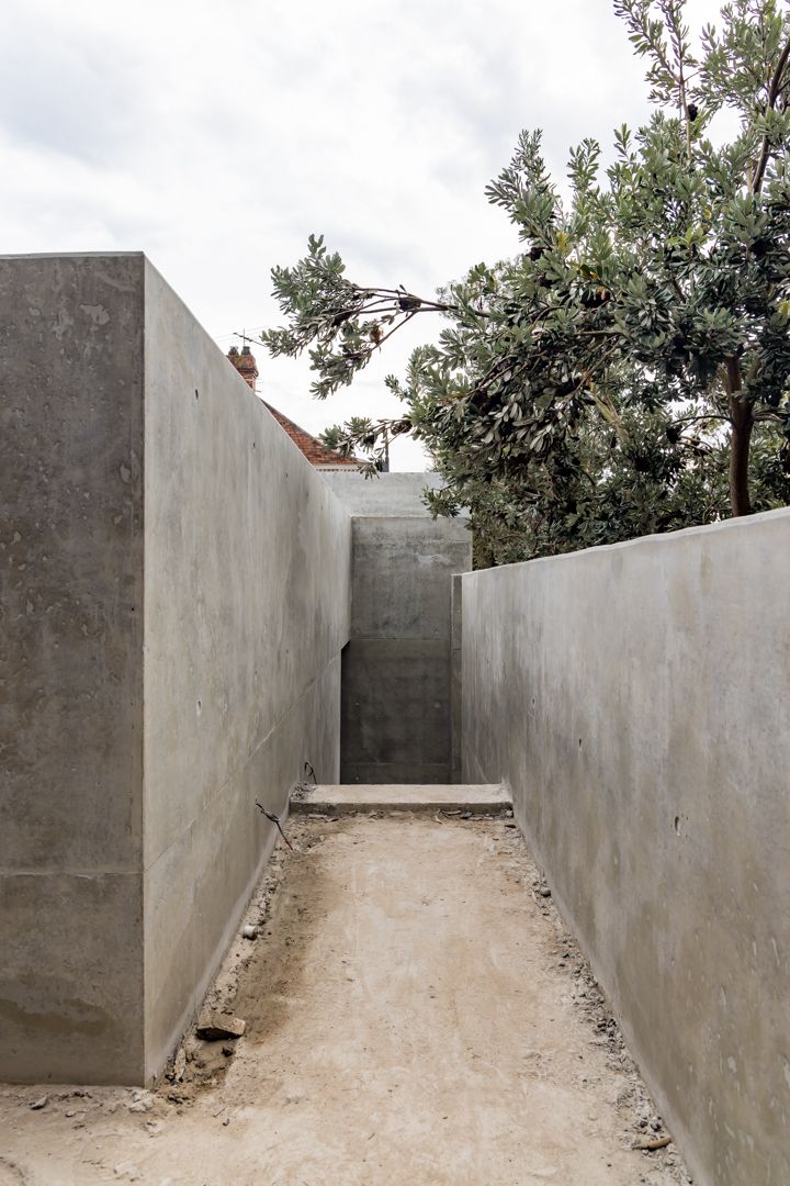 Narrow concrete passage between tall grey walls with a gravel floor and a tree overhanging against a cloudy sky.