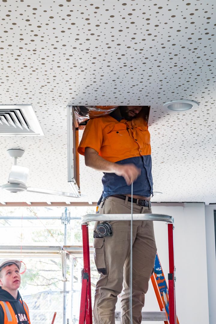 Electrician in an orange work shirt standing on a platform ladder inspecting wiring through an open ceiling hatch while a colleague watches.