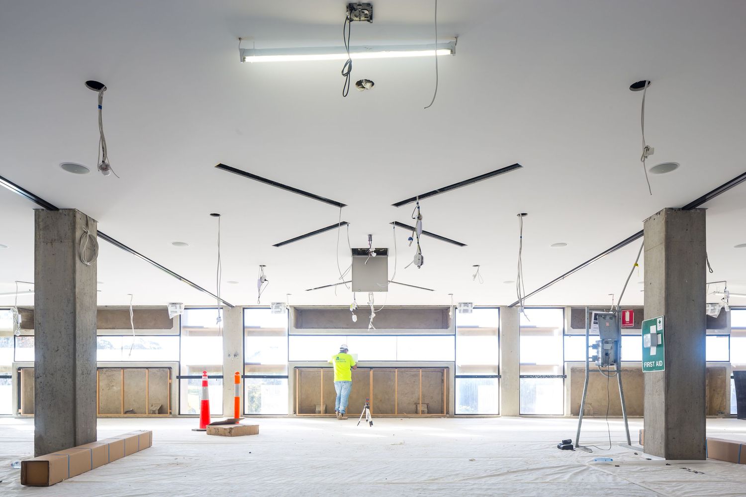Open concrete floor of a building under construction with columns, exposed ceiling services, traffic cones and a worker in hi-vis.