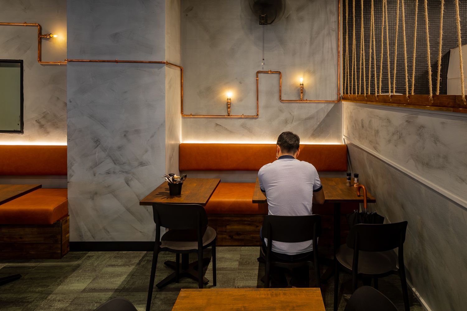 Lone diner seated at a wooden table in a moody restaurant with orange banquettes, marble walls and copper pipe lighting.