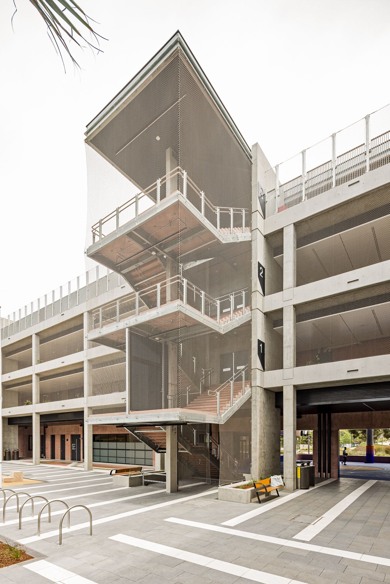 Modern concrete parking structure with an angular canopy over open staircases, steel railings and a bench on a paved plaza.