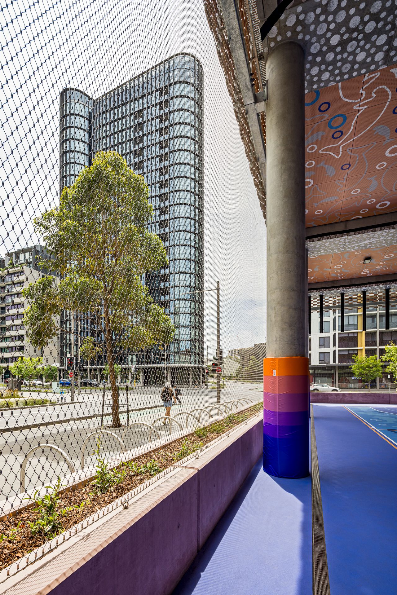 Rainbow-striped column and blue court floor beside mesh fencing, with a glass skyscraper and street trees in the background.