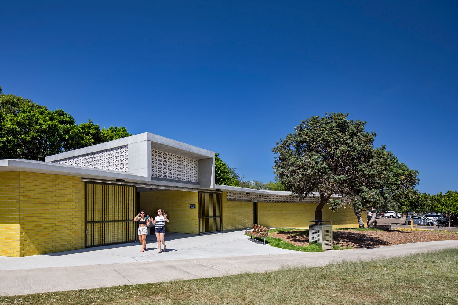 Two people walk past a yellow brick park amenities building with a concrete breeze block roof feature under a clear blue sky.