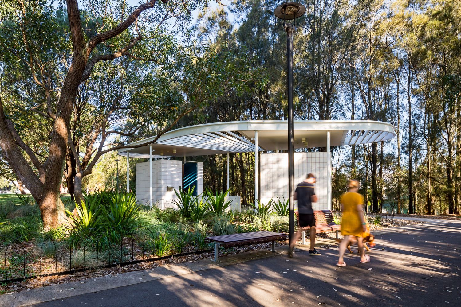 Two people walk along a park path past a bench and lamp post toward a white curved-roof amenities pavilion among trees.