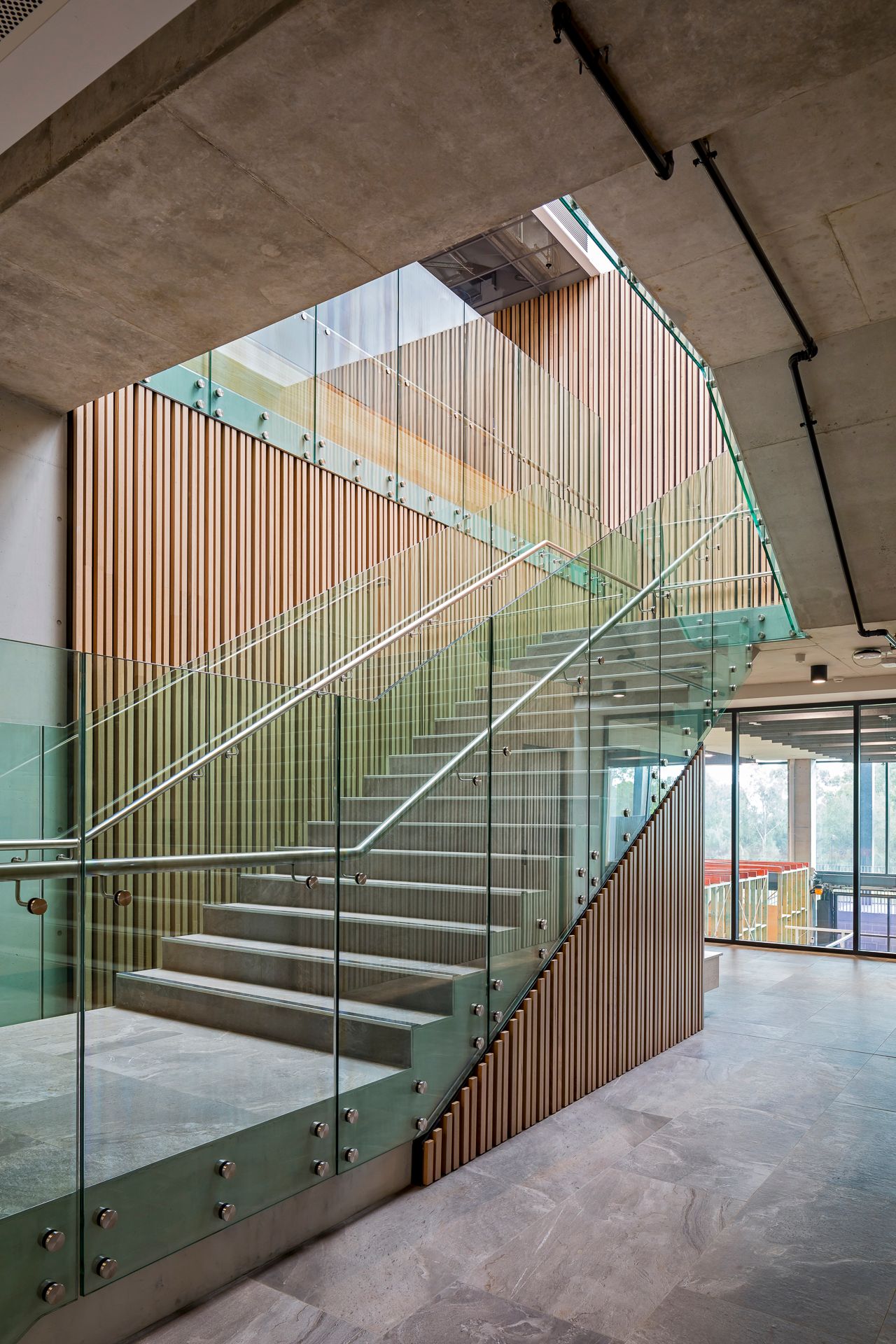 Contemporary glass-balustrade staircase with vertical timber battens and exposed concrete ceiling in a modern campus building interior.