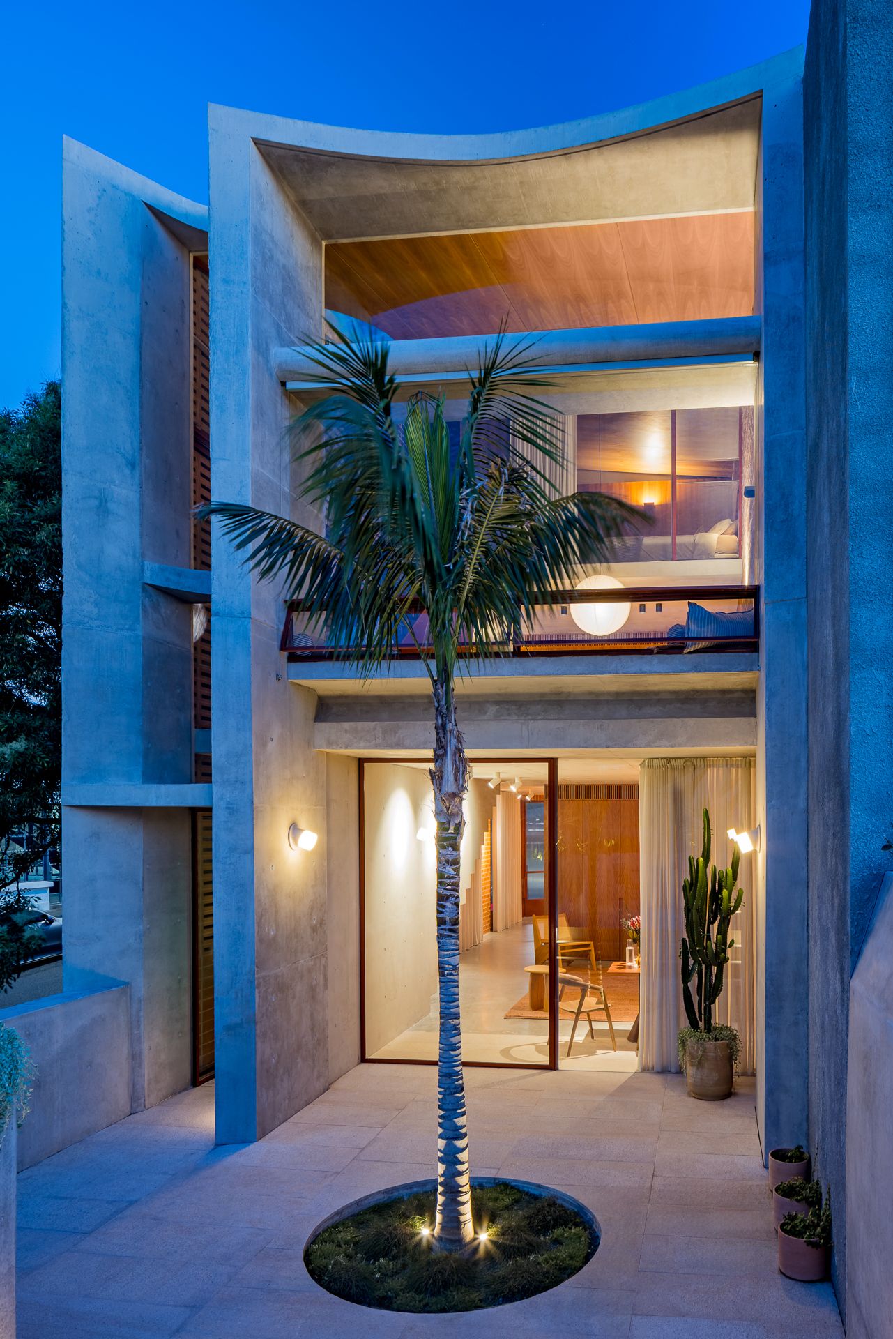 Contemporary concrete townhouse facade with curved balcony walls, glowing interiors, and a palm tree in the courtyard at dusk.