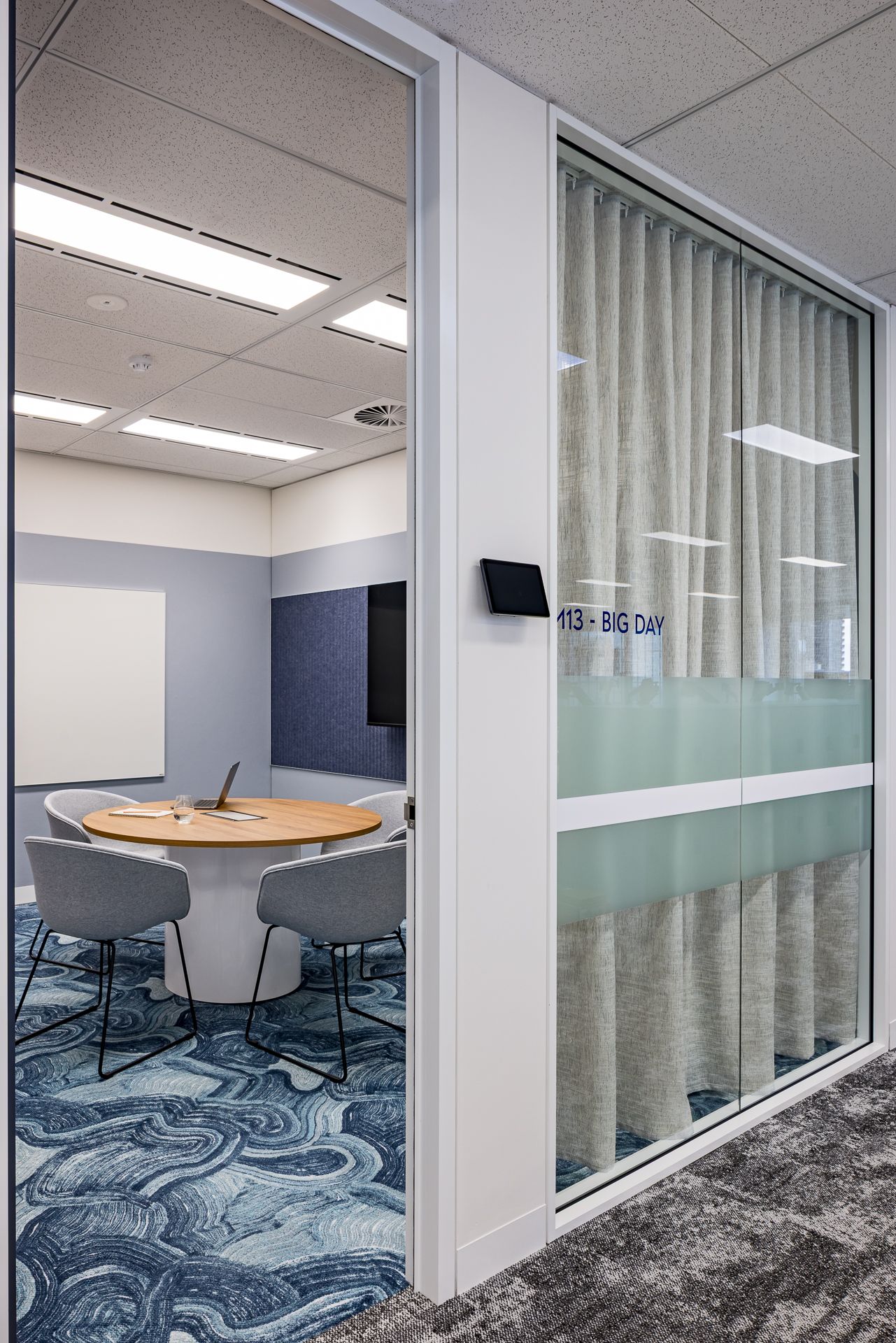 Glass-fronted meeting room labeled Big Day, with a round table, gray chairs, blue wave-patterned carpet and a privacy curtain.