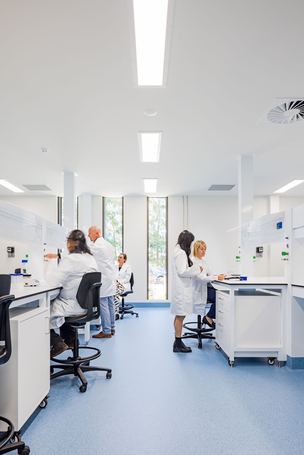 Team of scientists in lab coats working at benches in a spacious white laboratory with blue flooring and views of the trees outside.