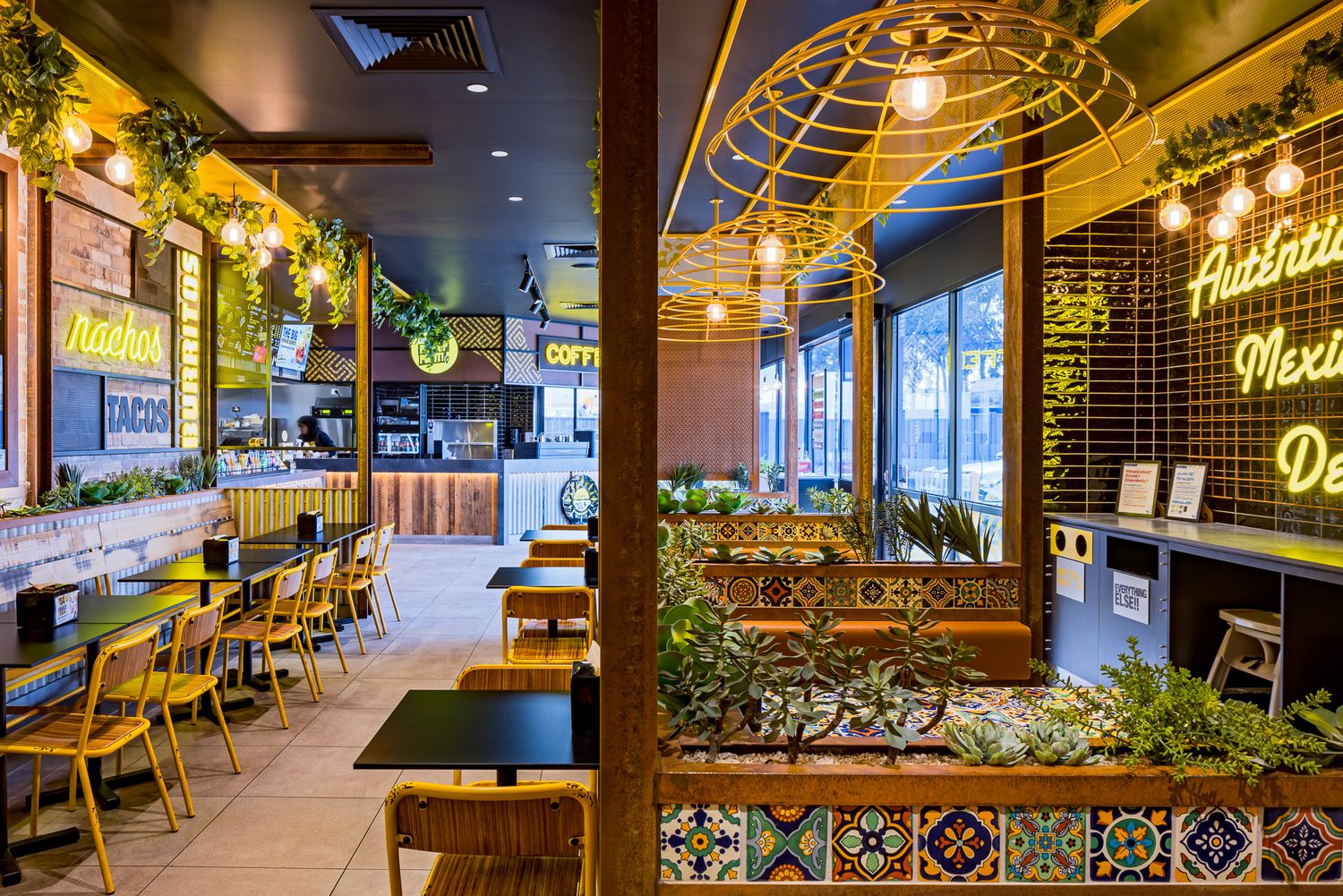 Bright restaurant interior with neon Mexican signs, yellow chairs, tiled planters and warm string lighting over the dining area.