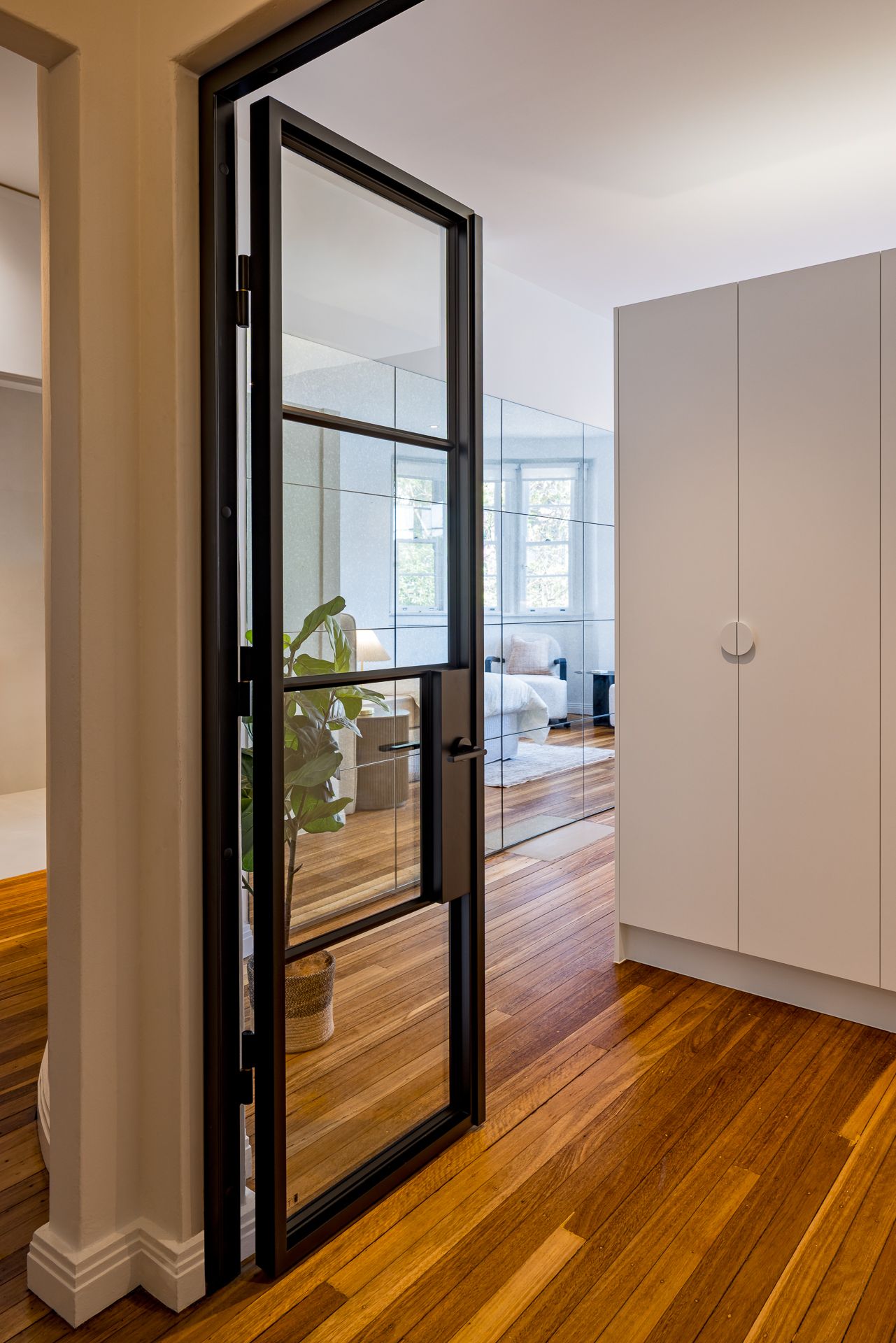 Steel-framed glass door with a potted plant leads to a bedroom beside a white wardrobe on a timber floor.