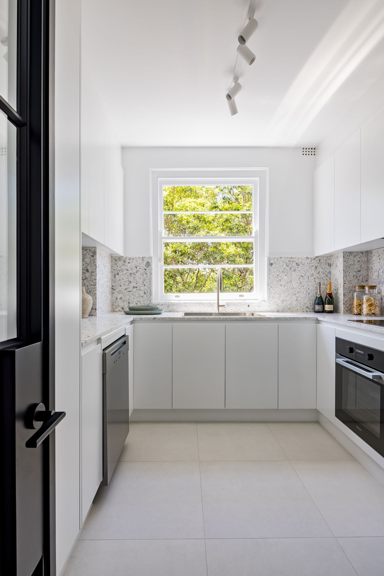 Galley-style white kitchen with stone benchtops, stainless dishwasher, built-in oven, track lighting and a sunny garden window.