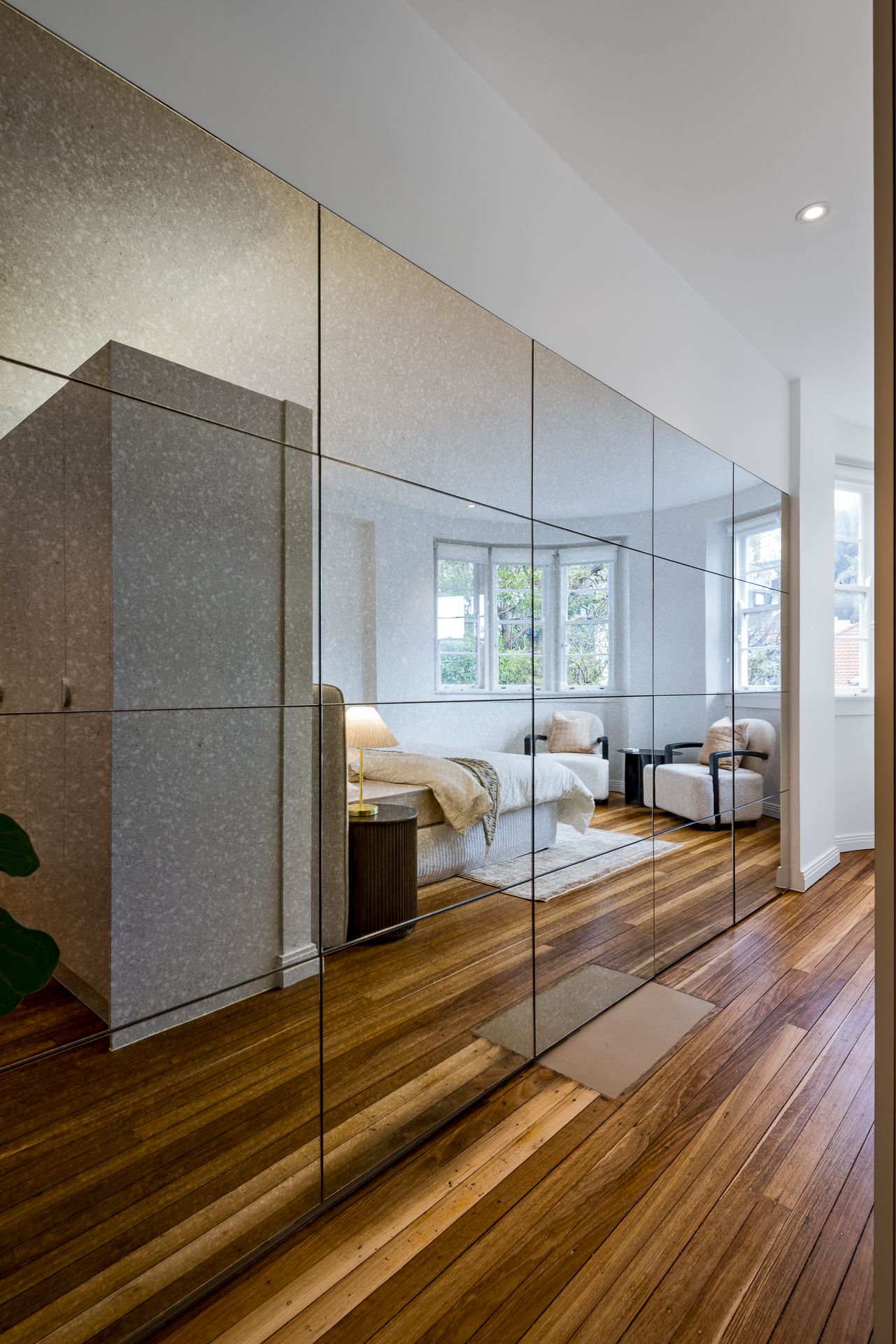 Mirrored wall panels in a timber-floored hallway reflecting a bright bedroom with a bed, bedside lamp and a cozy armchair.