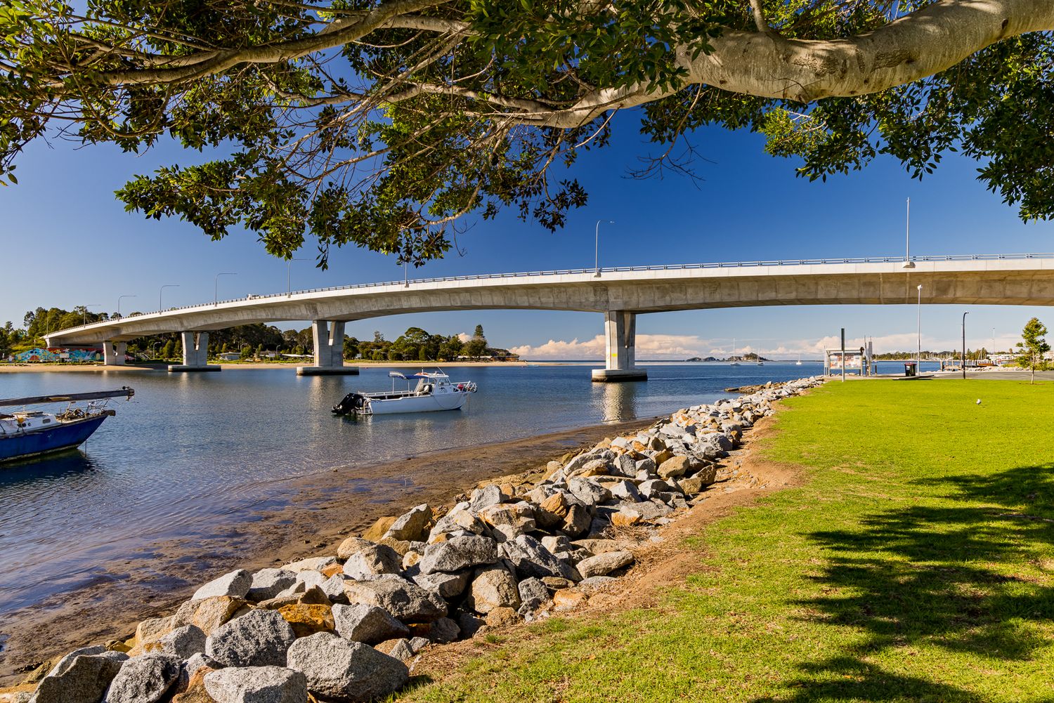 Rock-lined shoreline and grassy park beside a calm bay with moored boats and a long curved bridge in the background.