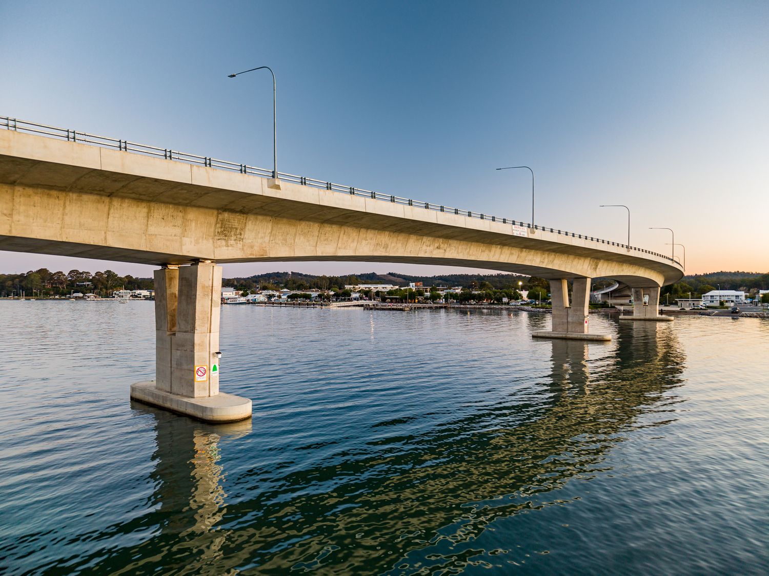 Concrete waterway bridge with street lamps reflected in still water under a soft sunset sky.