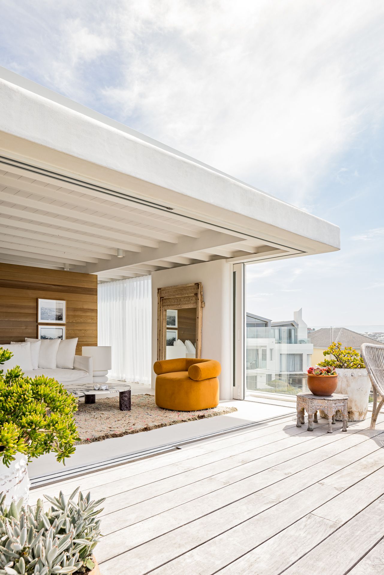 Beachfront terrace with weathered timber decking, mustard armchair, white sofa and succulents beside a covered living area.
