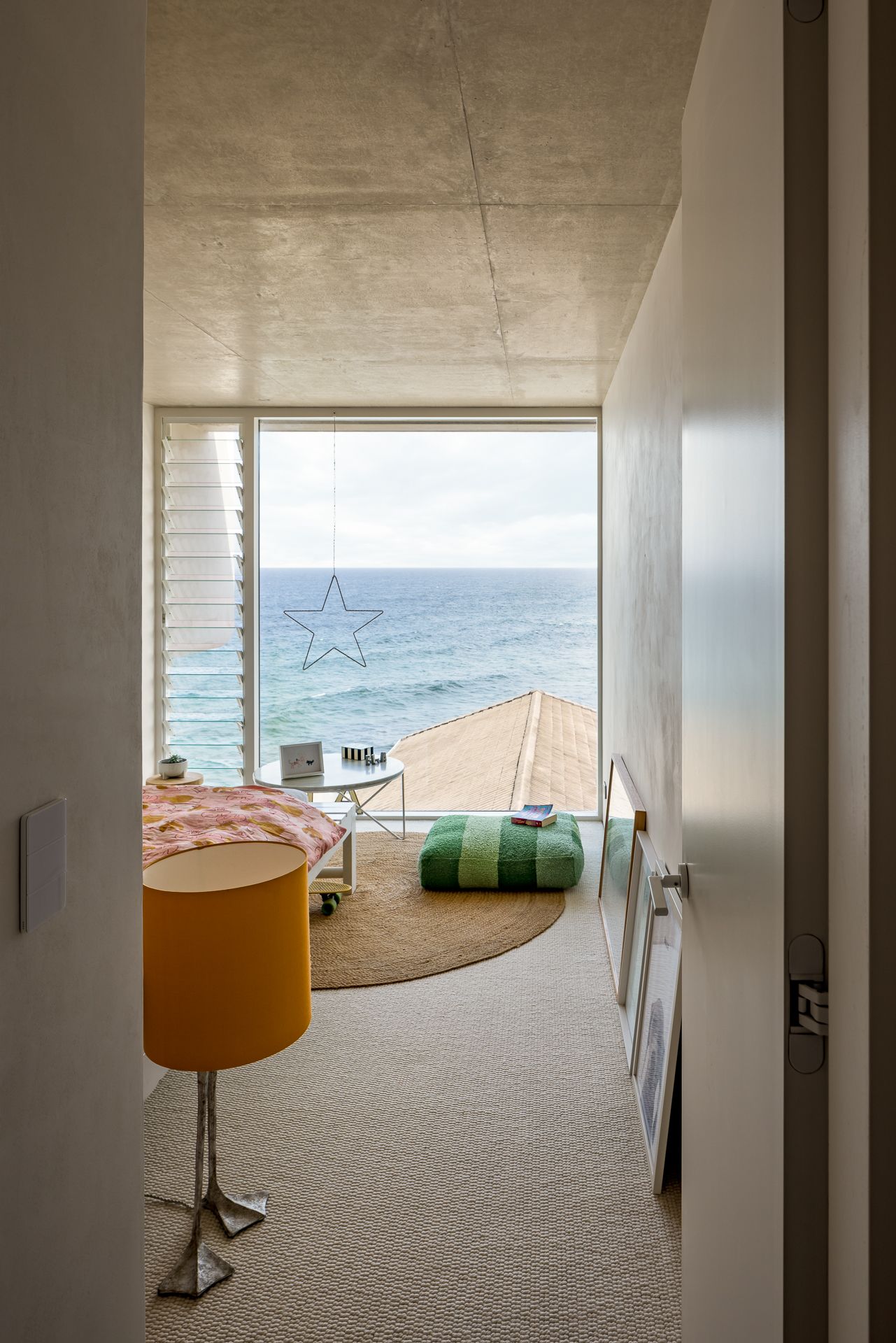 Bedroom with a concrete ceiling and floor-to-ceiling window framing ocean views, with a green floor cushion and orange lamp.