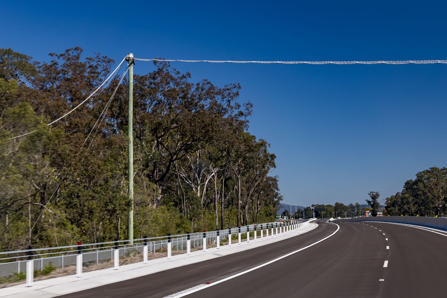 Curving new asphalt road with white guardrail posts beside a power pole and eucalyptus trees under a bright blue sky.