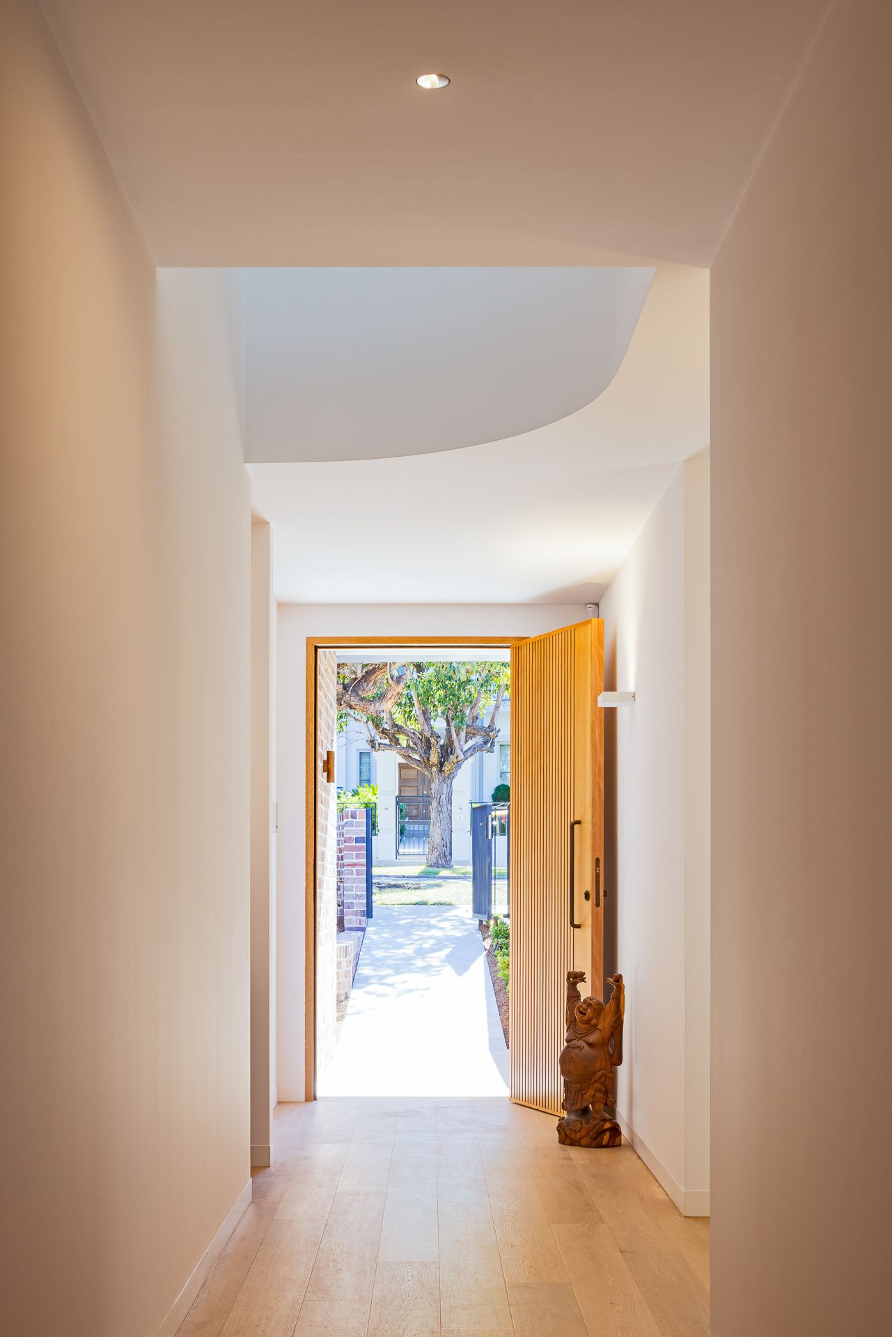 Light-filled hallway with timber flooring and a curved ceiling leading to an open wooden front door and a carved timber figure.