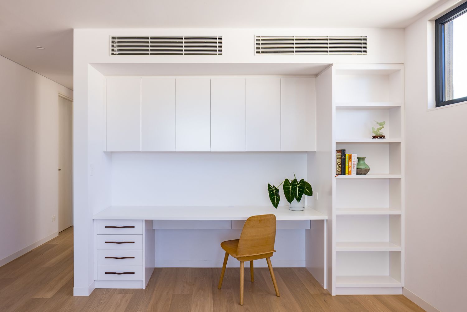 Built-in white study nook with desk, drawers, overhead cabinets and open shelves beside a wooden chair on oak flooring.