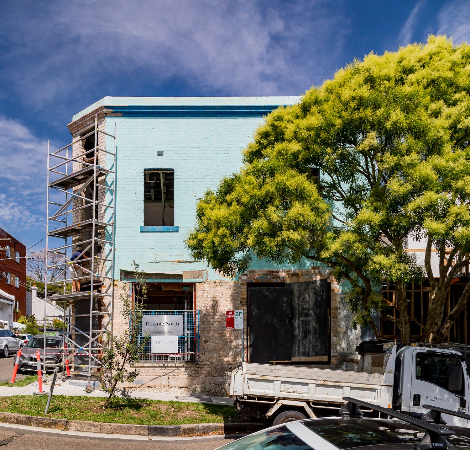 Heritage corner building with pale blue facade and scaffolding, framed by a flowering tree with a work truck parked nearby.