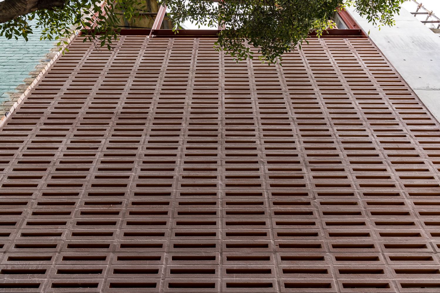 Upward view of a tall building facade made of terracotta-colored perforated brick screens, with tree branches at the top left corner.
