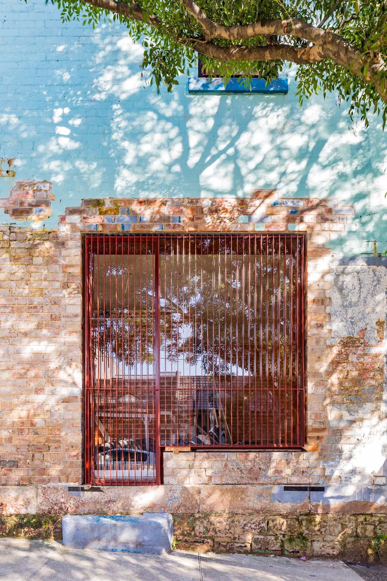 Old brick wall with a large square window covered by red vertical slats and dappled tree shadows.