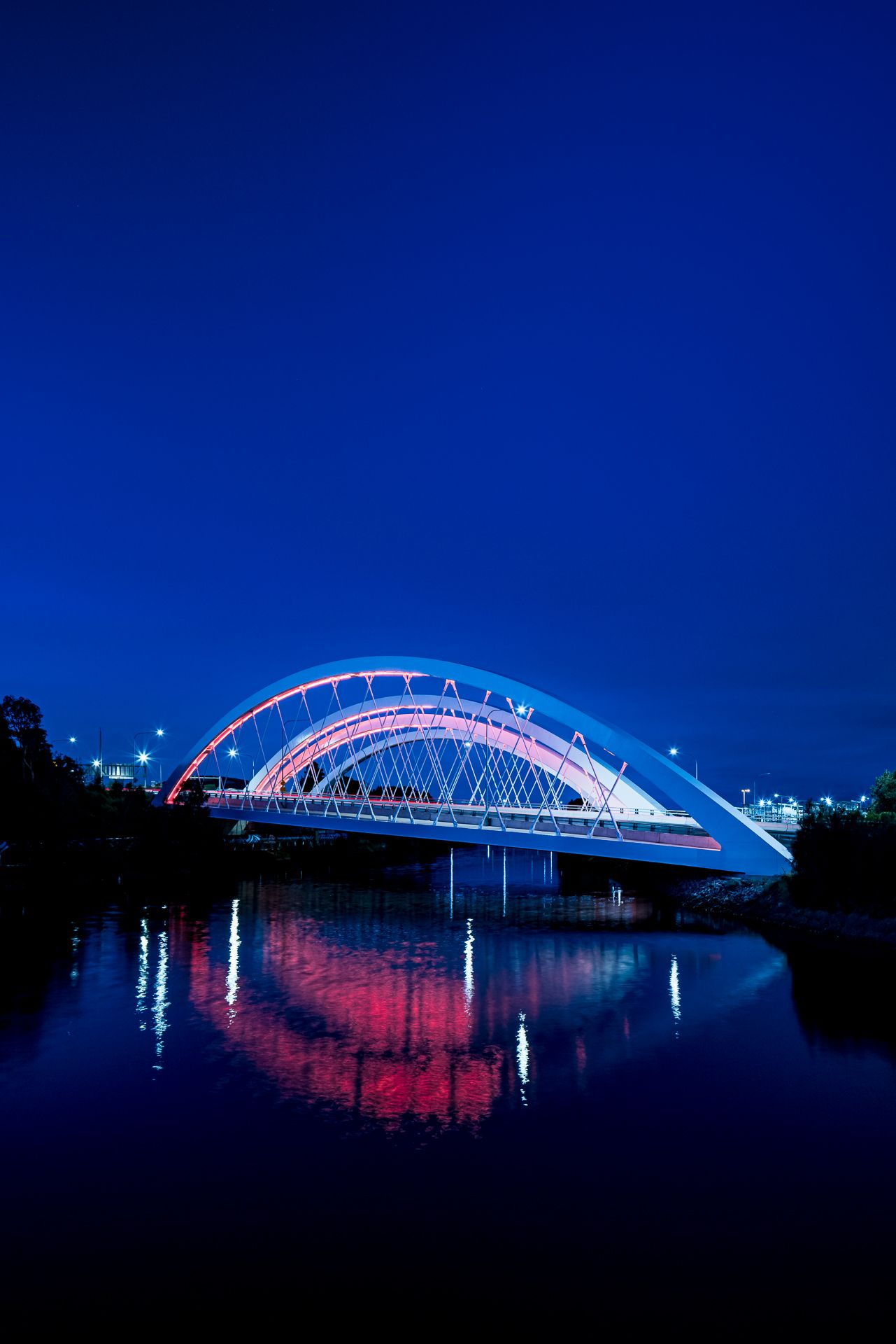 Illuminated arch bridge with pink and red lights reflected in a calm river beneath a deep blue night sky.