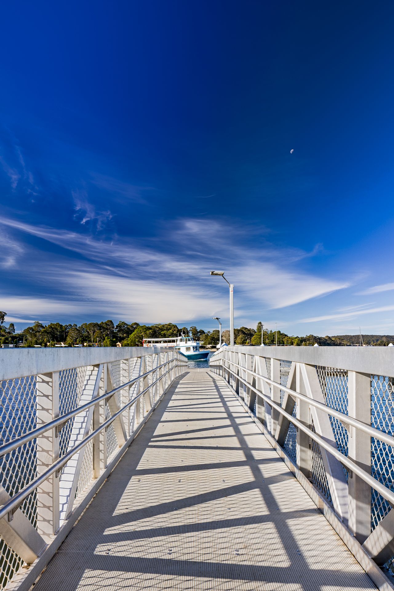 White metal pedestrian jetty walkway with railings leading toward a boat on the water under a deep blue sky with wispy clouds.