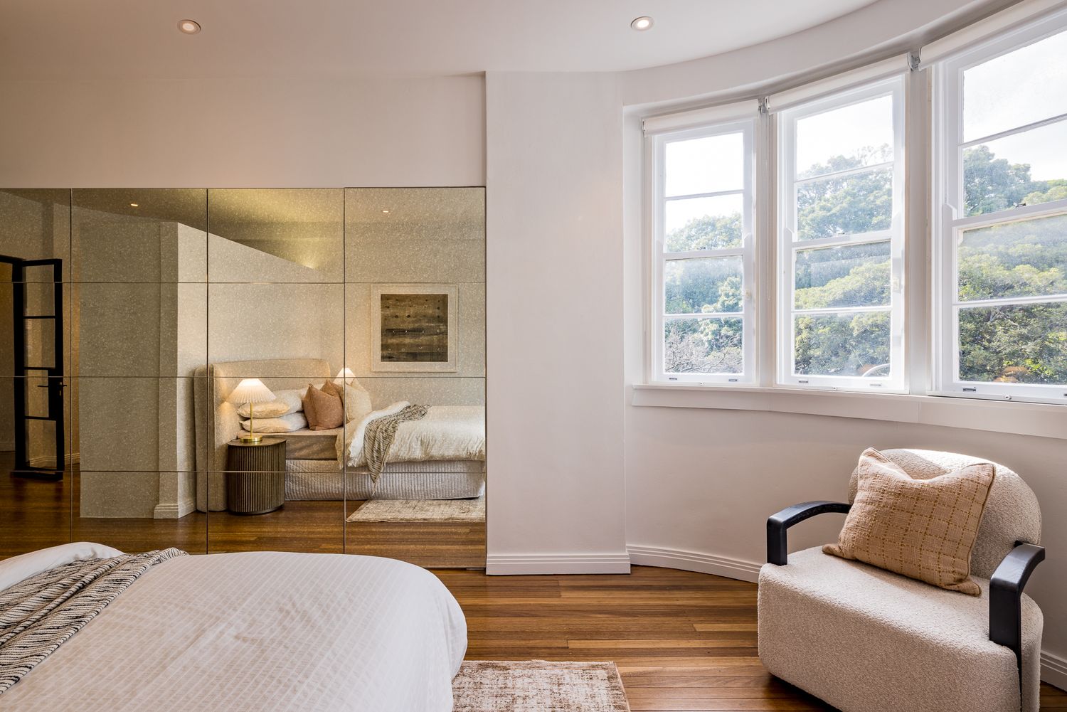 Sunlit bedroom with curved bay windows overlooking trees, a cushioned armchair, mirrored wardrobe and polished timber floors.