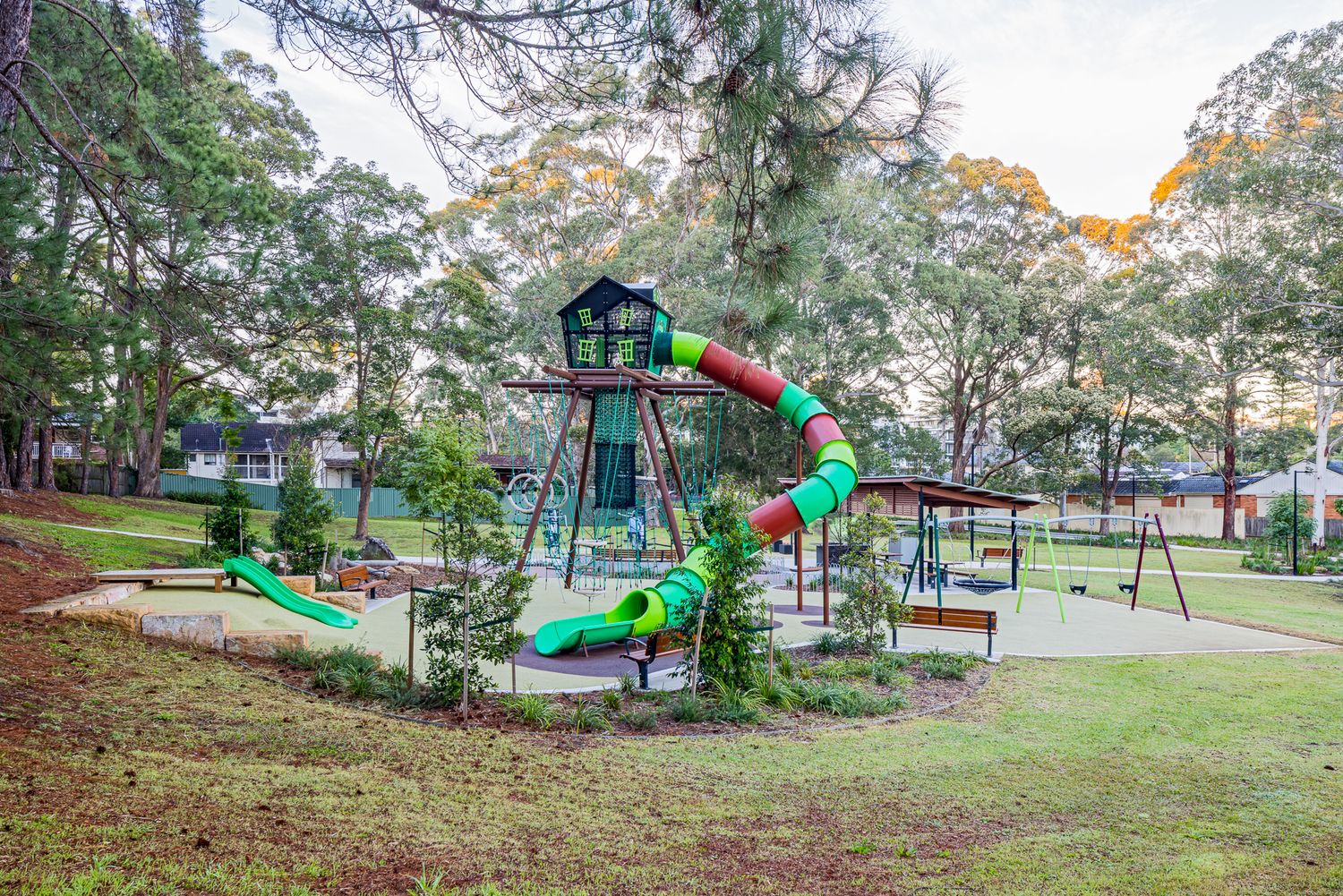 Wide view of a park playground with a spiral tube slide, climbing tower, shade shelter and benches beside a grassy lawn and tall trees.