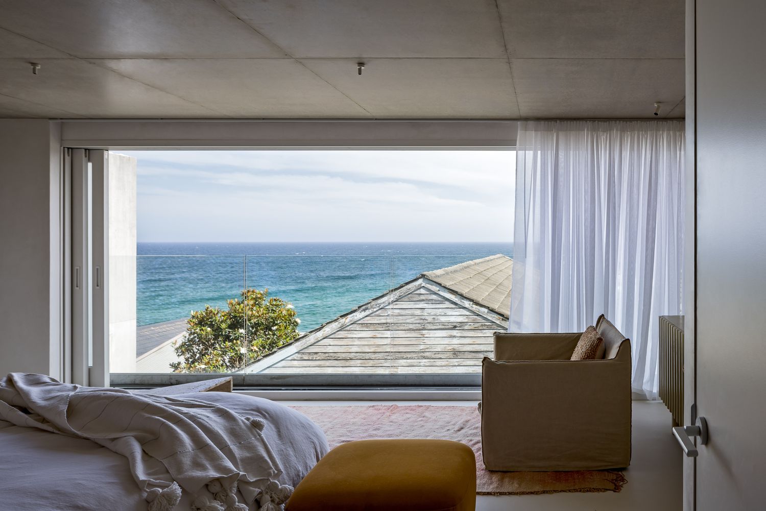 Bedroom with floor-to-ceiling windows framing a sweeping ocean view, a cream armchair, sheer curtains and an ochre ottoman.