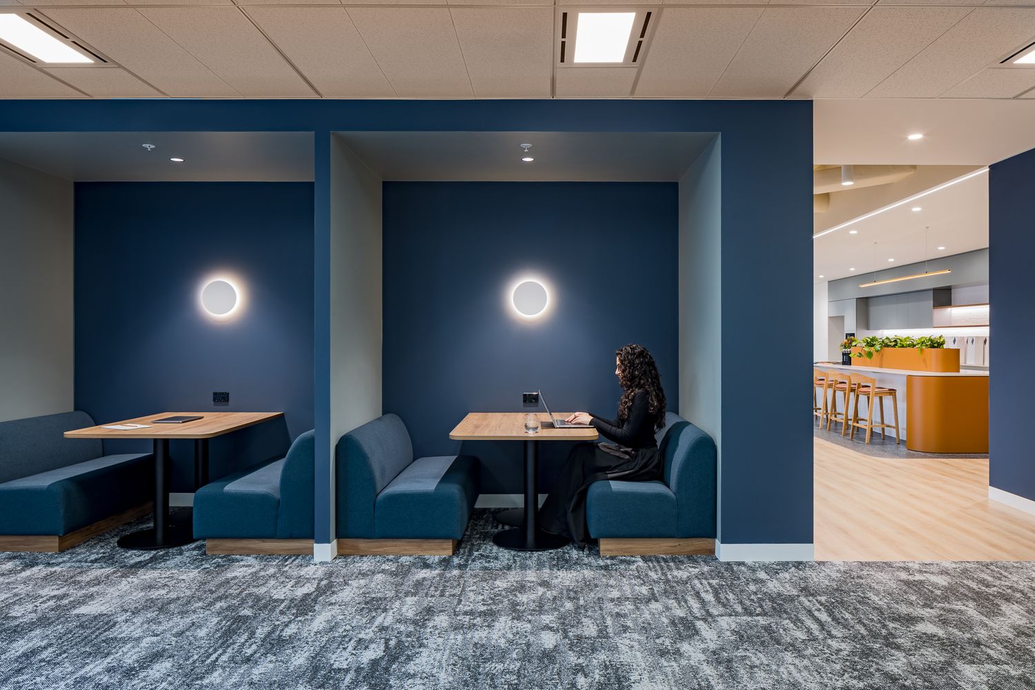 Woman working on a laptop in a navy blue booth in a modern office, with a kitchen and breakout area visible in the background.