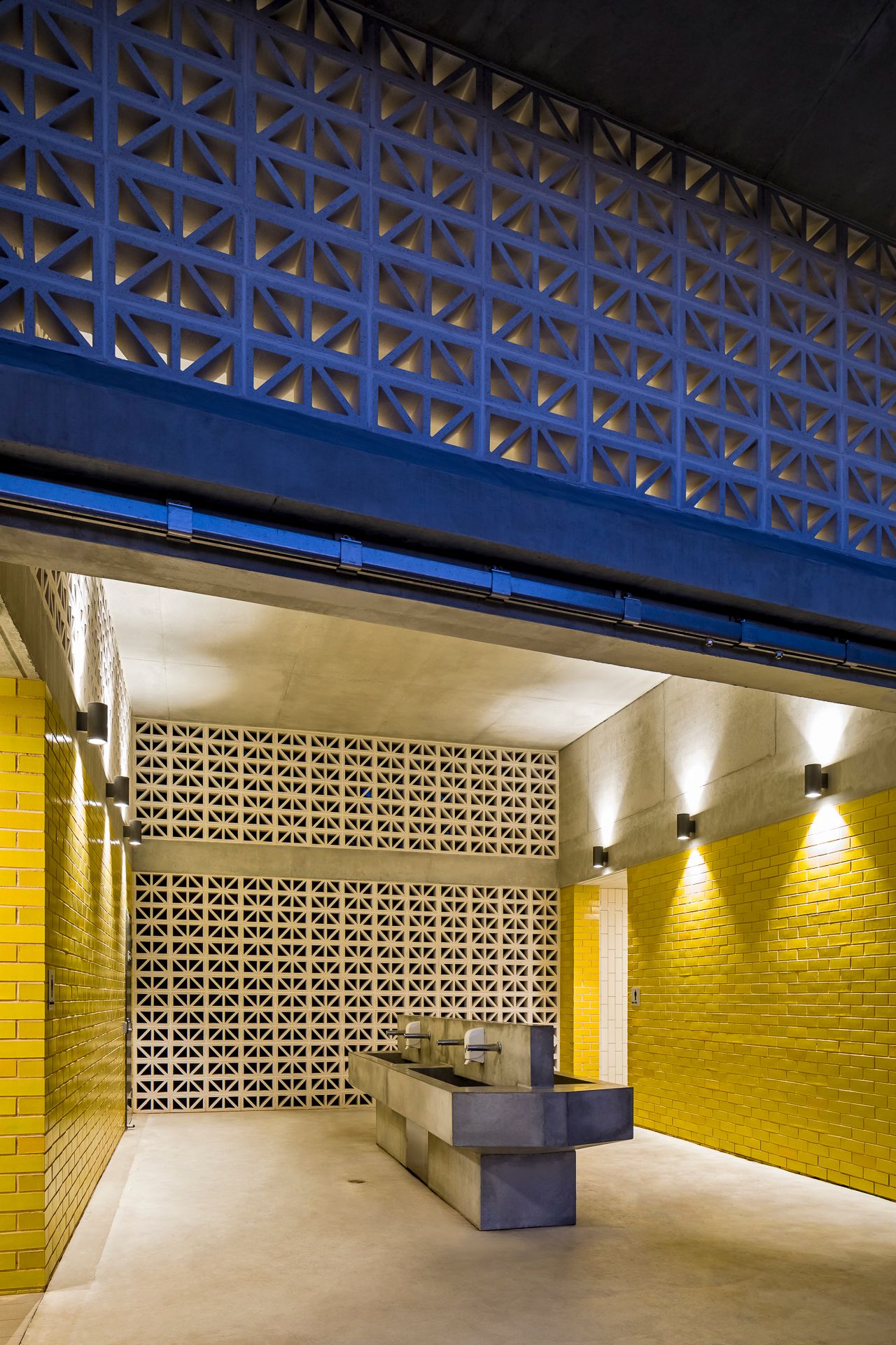 Public restroom interior with yellow brick walls, blue breeze block screen above, and a concrete trough sink lit by wall lights.