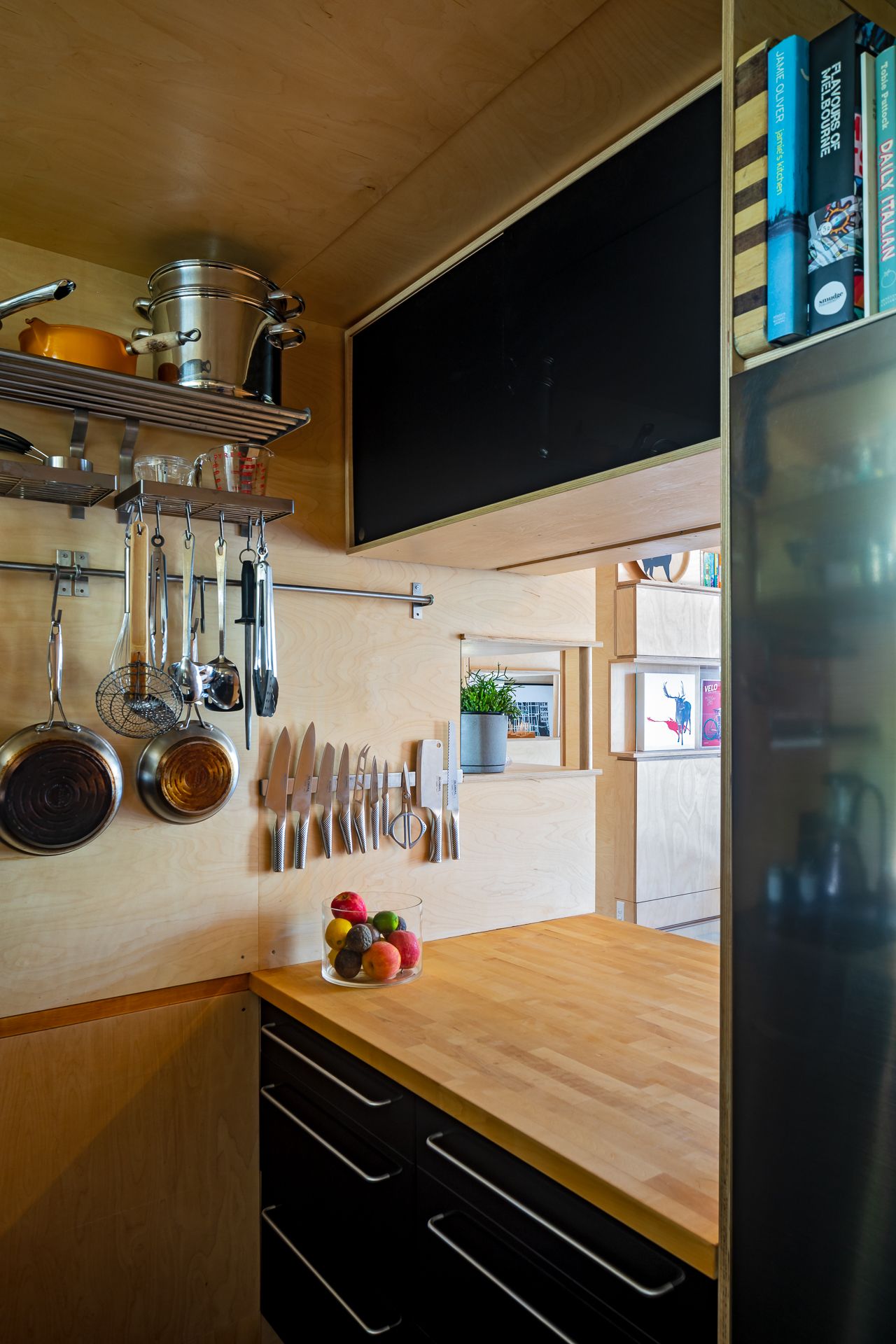 Compact kitchen with butcher block counter, black cabinets, hanging pots and knives, and a bowl of fresh fruit.