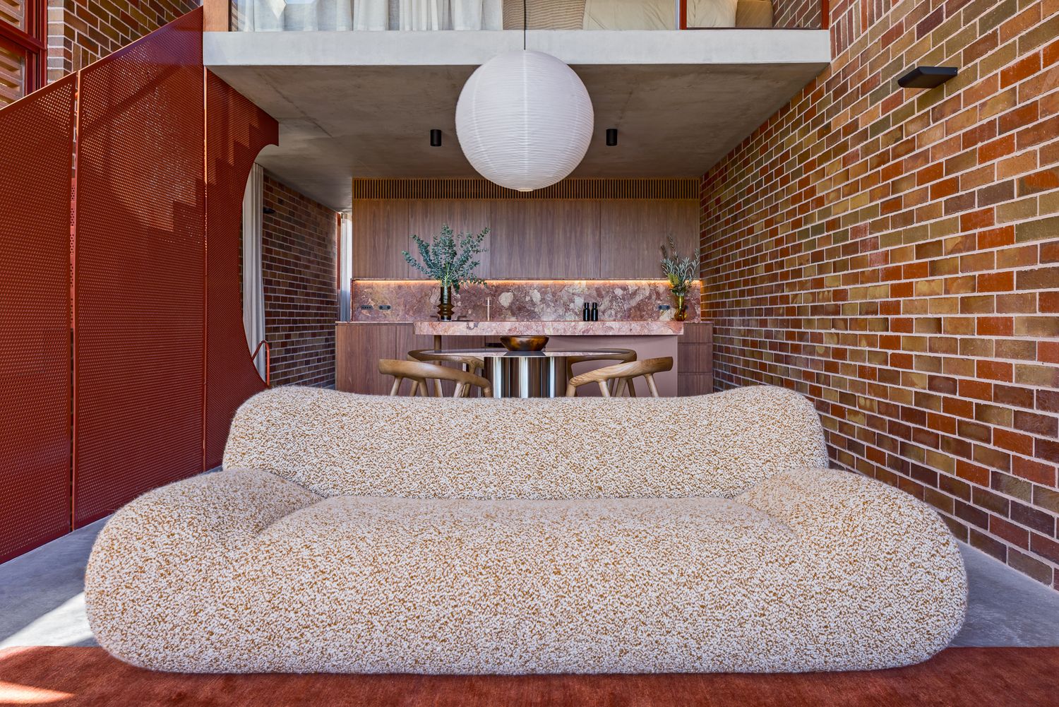 Textured boucle sofa in a brick-walled living space with a red perforated screen, pendant lamp and dining area beyond.