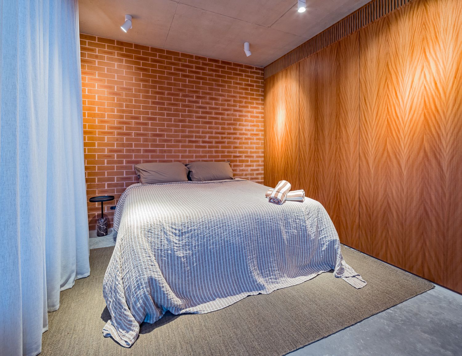Warm bedroom with a white bed, rolled towels, exposed orange brick wall, grain timber paneling and sheer curtains on a concrete floor.