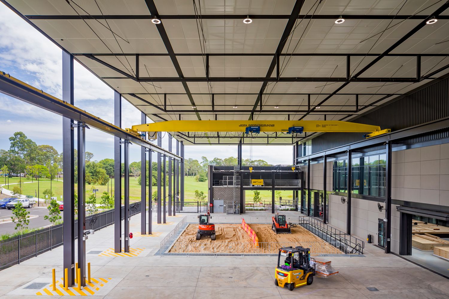 Spacious workshop with glass walls, overhead yellow gantry crane, and small construction vehicles on a sand training pit with garden views.