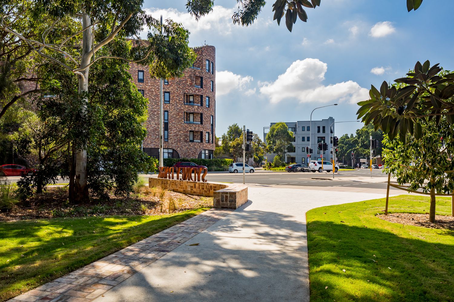 Park pathway with a Sydney Park sign leading toward brick apartment buildings, framed by trees and green lawn.