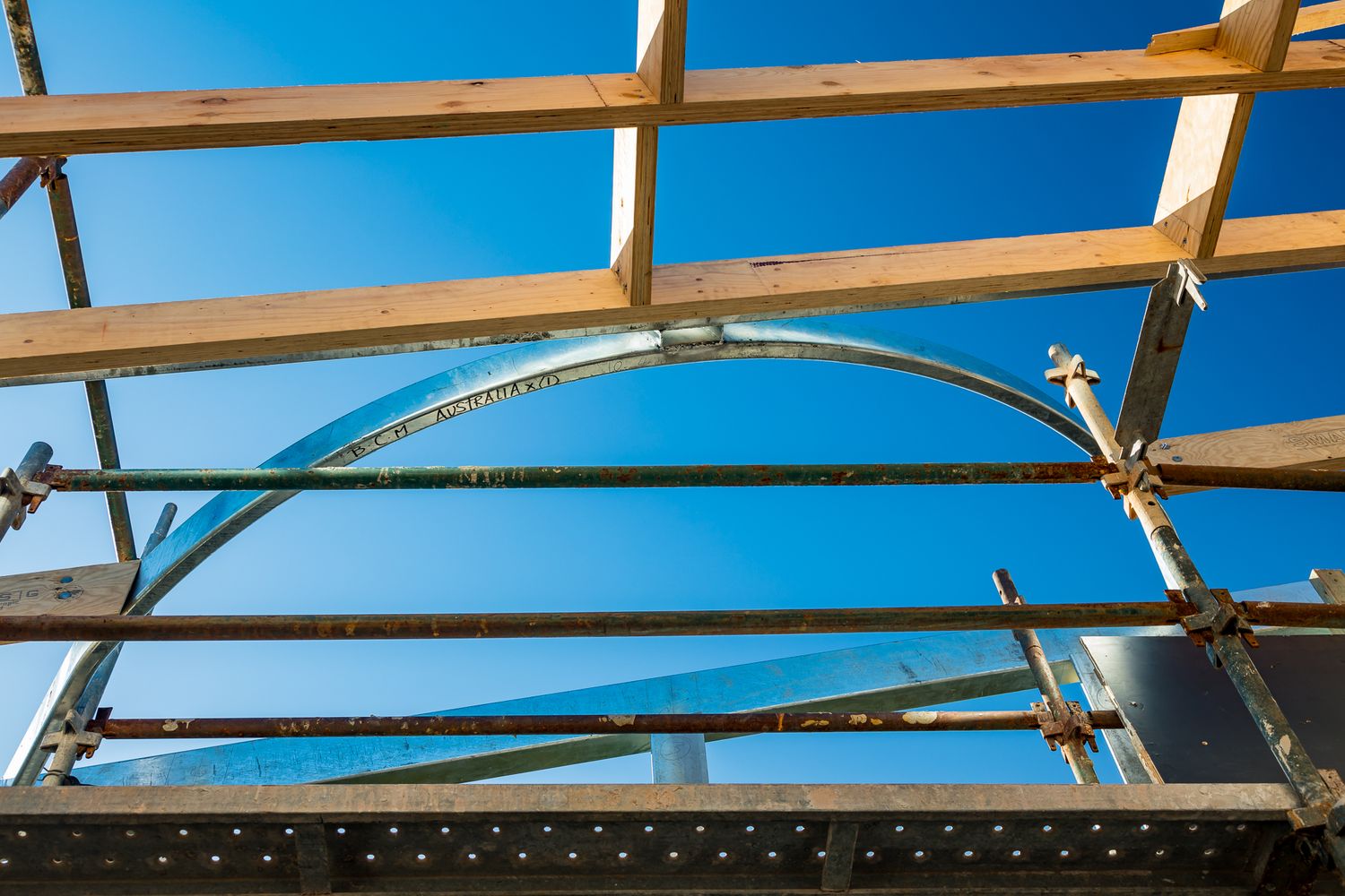 Timber roof battens and scaffolding poles frame a curved steel arch against a bright blue sky at a building site.