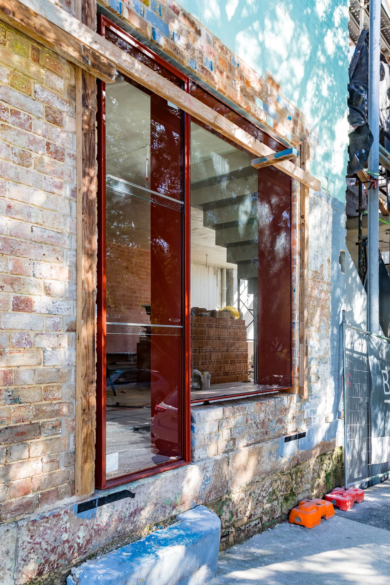 Red-framed shopfront window in a renovated old brick building with a stone sill and footpath outside.