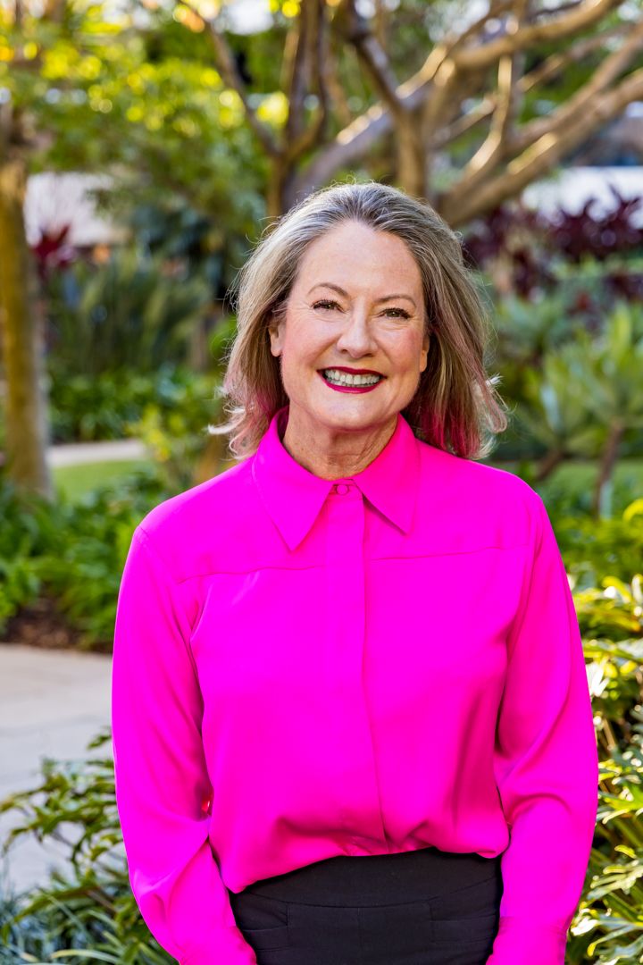 Smiling woman with silver-gray hair and a bright pink blouse standing outdoors in front of lush green garden foliage.