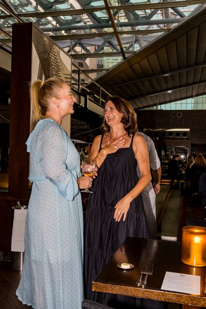 Two smiling women in long dresses chat and hold drinks at an event in a modern venue with a glass ceiling.