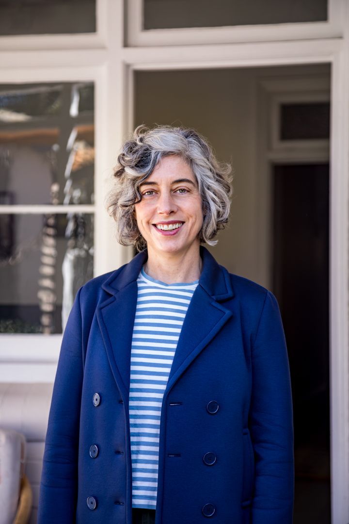 Smiling woman with curly gray hair wearing a navy double-breasted coat and striped top stands outside a building with large windows.