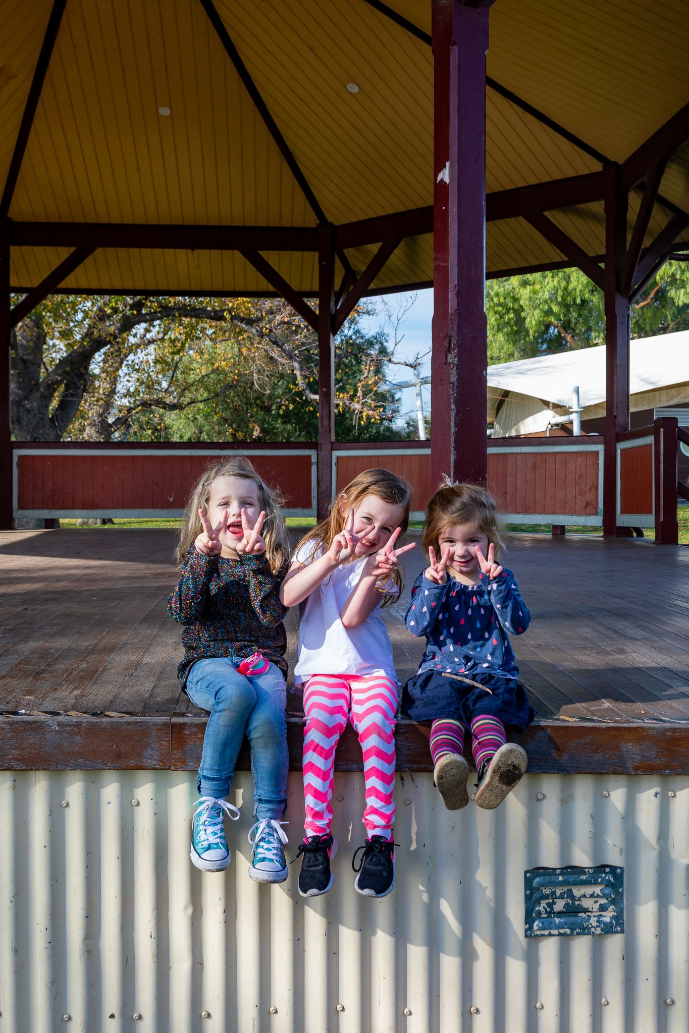 Three young girls sit side by side on the edge of a wooden gazebo platform, smiling and making playful hand gestures for the camera.