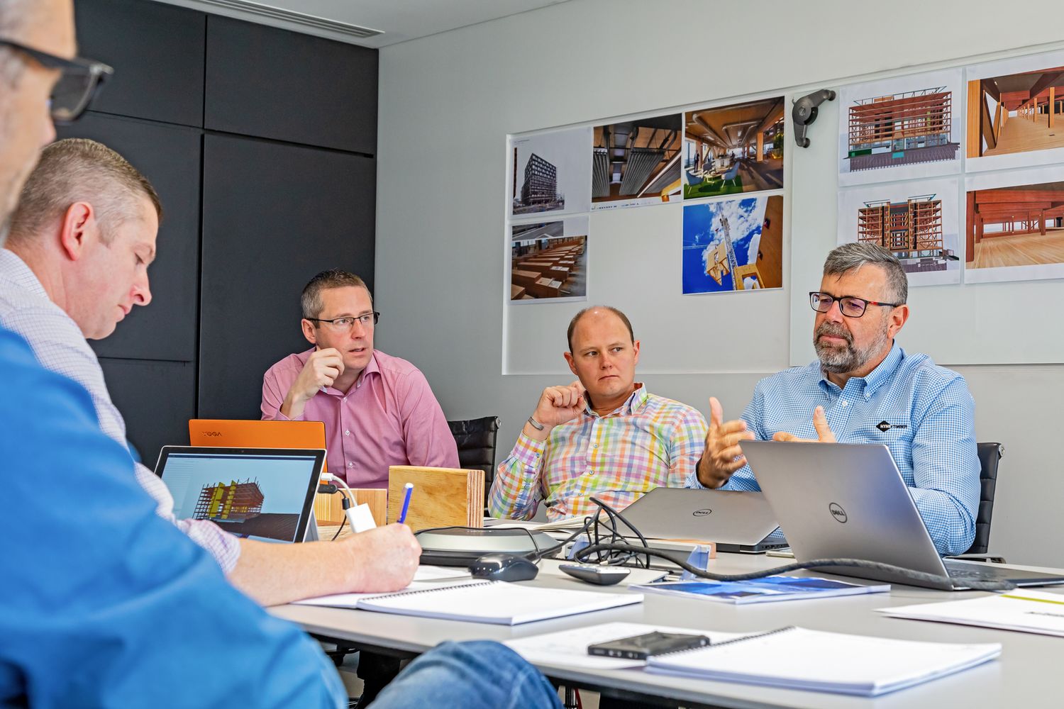 Four professionals hold a business meeting around a conference table with laptops and documents, with architectural photos on the wall behin